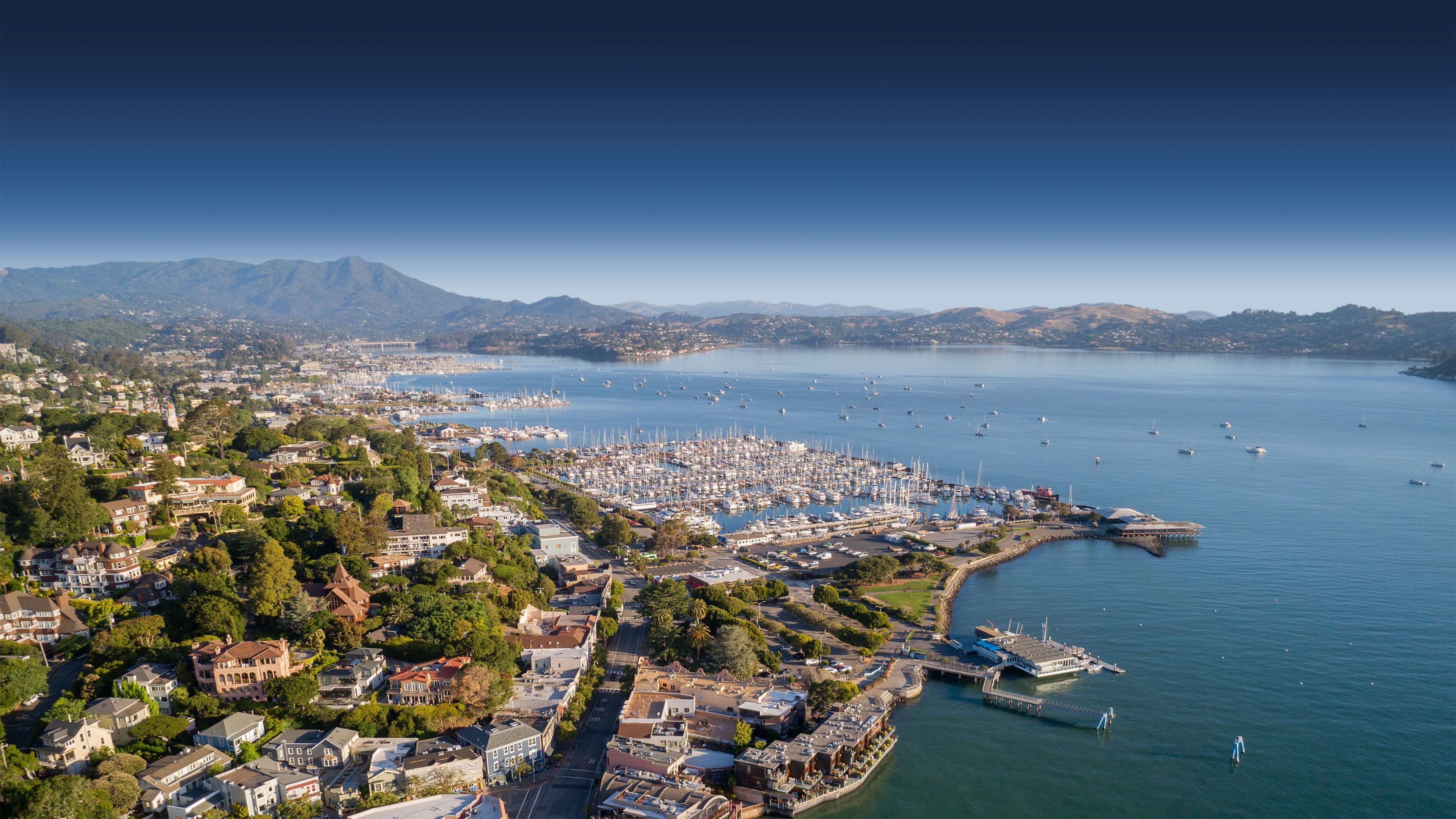 Aerial view of Sausalito, its waterfront, marinas, hills, and Richardson Bay beneath an open blue sky.