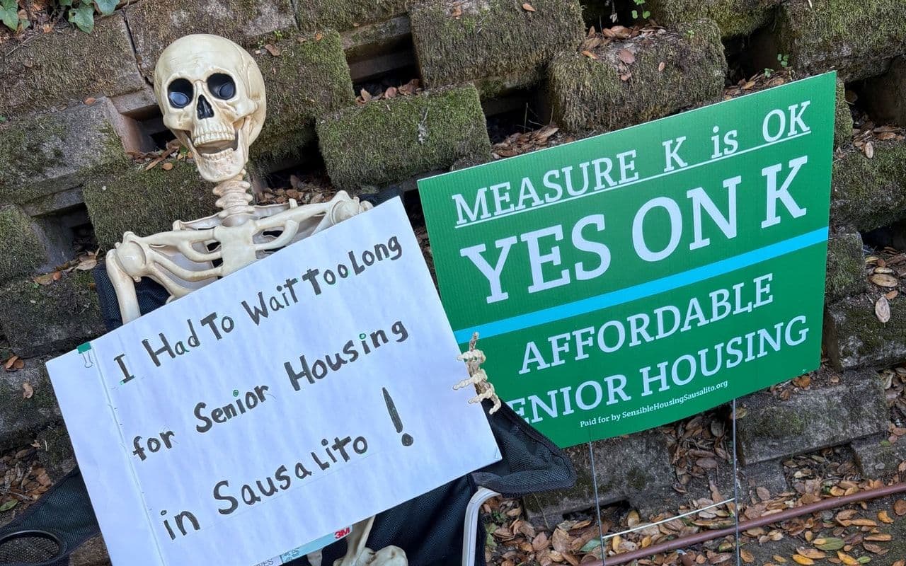 A skeleton figure seated outside holding a hand-lettered sign — 'I had to wait too long for senior housing in Sausalito' — beside a green 'Yes on K · Affordable Senior Housing' sign.